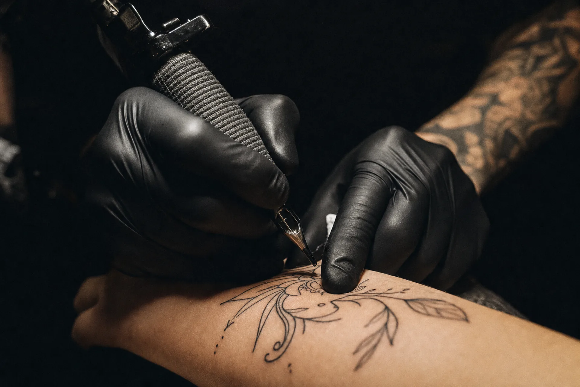 Mains gantées d'un tatoueur au-dessus d'un avant-bras portant un dessin au trait noir.